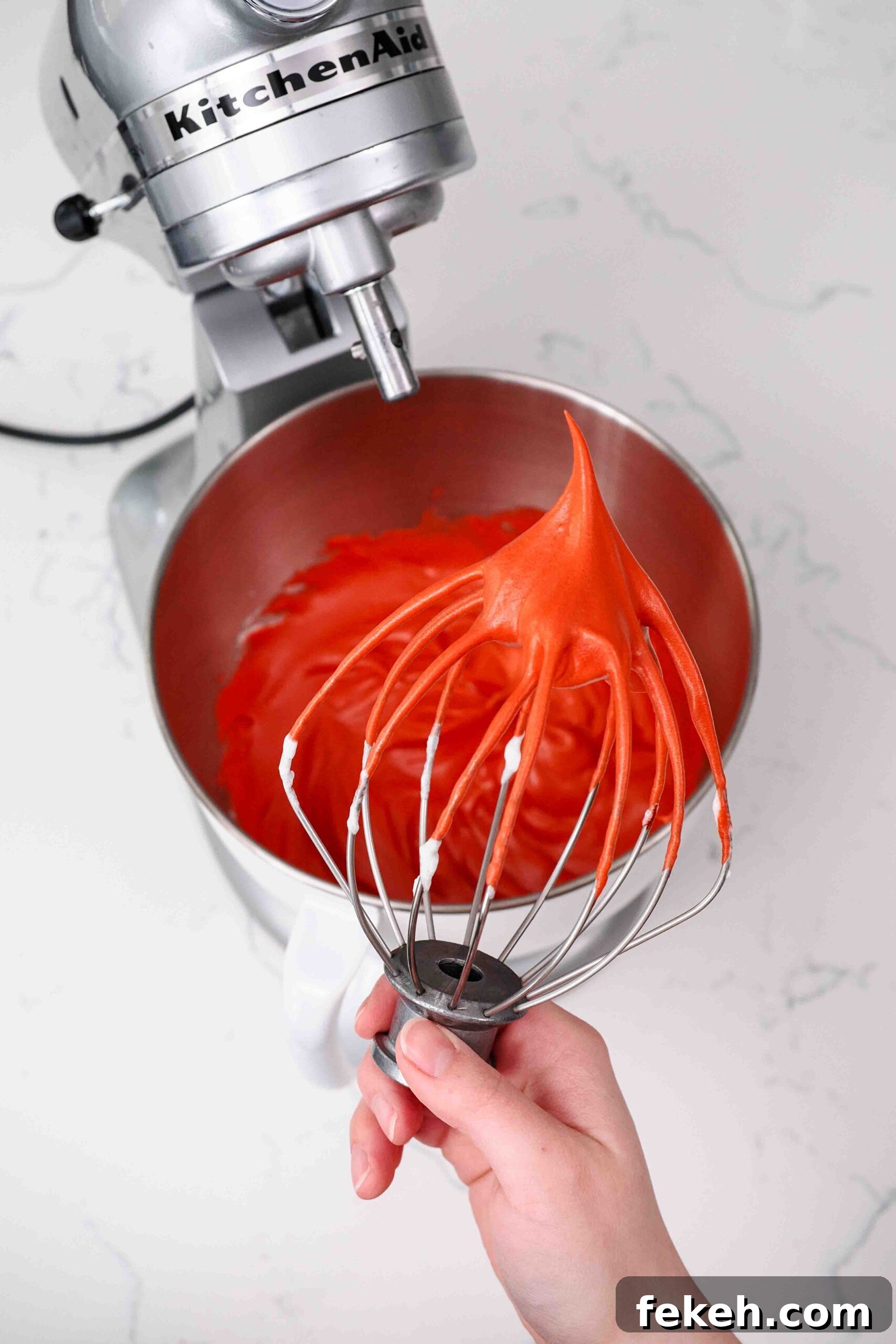 A hand holds up a whisk with a stiff peak of red meringue pointing up.