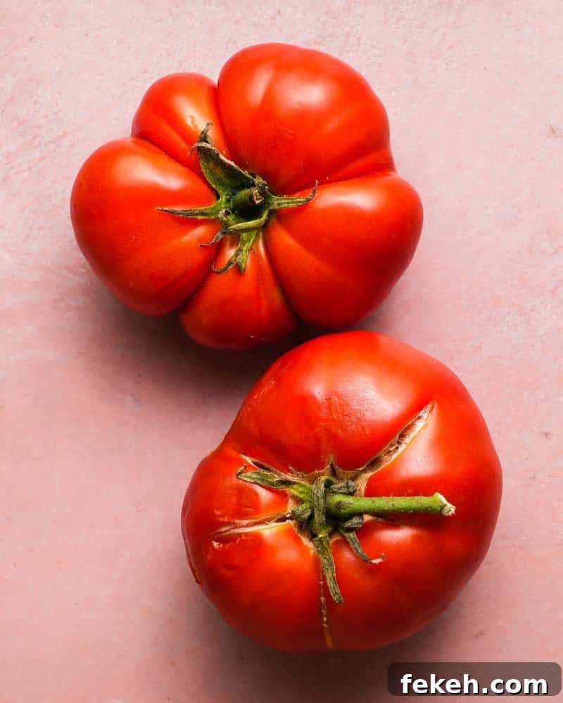 Creamy Vegan Roasted Tomato and Basil Soup Recipe 4 Two ripe red tomatoes with green stems on a light pink surface.