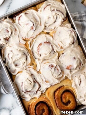 pan of brioche cinnamon rolls frosted except for 2 rolls with a kitchen towel underneath the pan