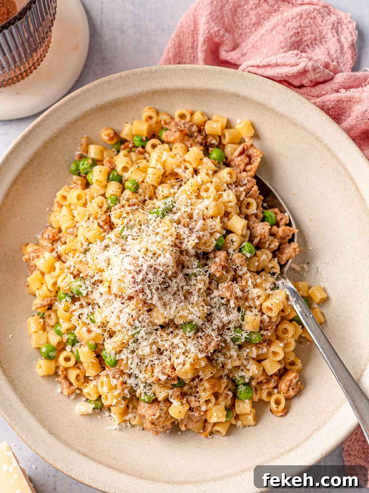 Ditalini Pasta with Italian Sausage and Peas 9 A bowl of ditalini with sausage and peas topped with a generous mound of grated Parmesan and black pepper, with a spoon in the bowl and a pink linen in the background.