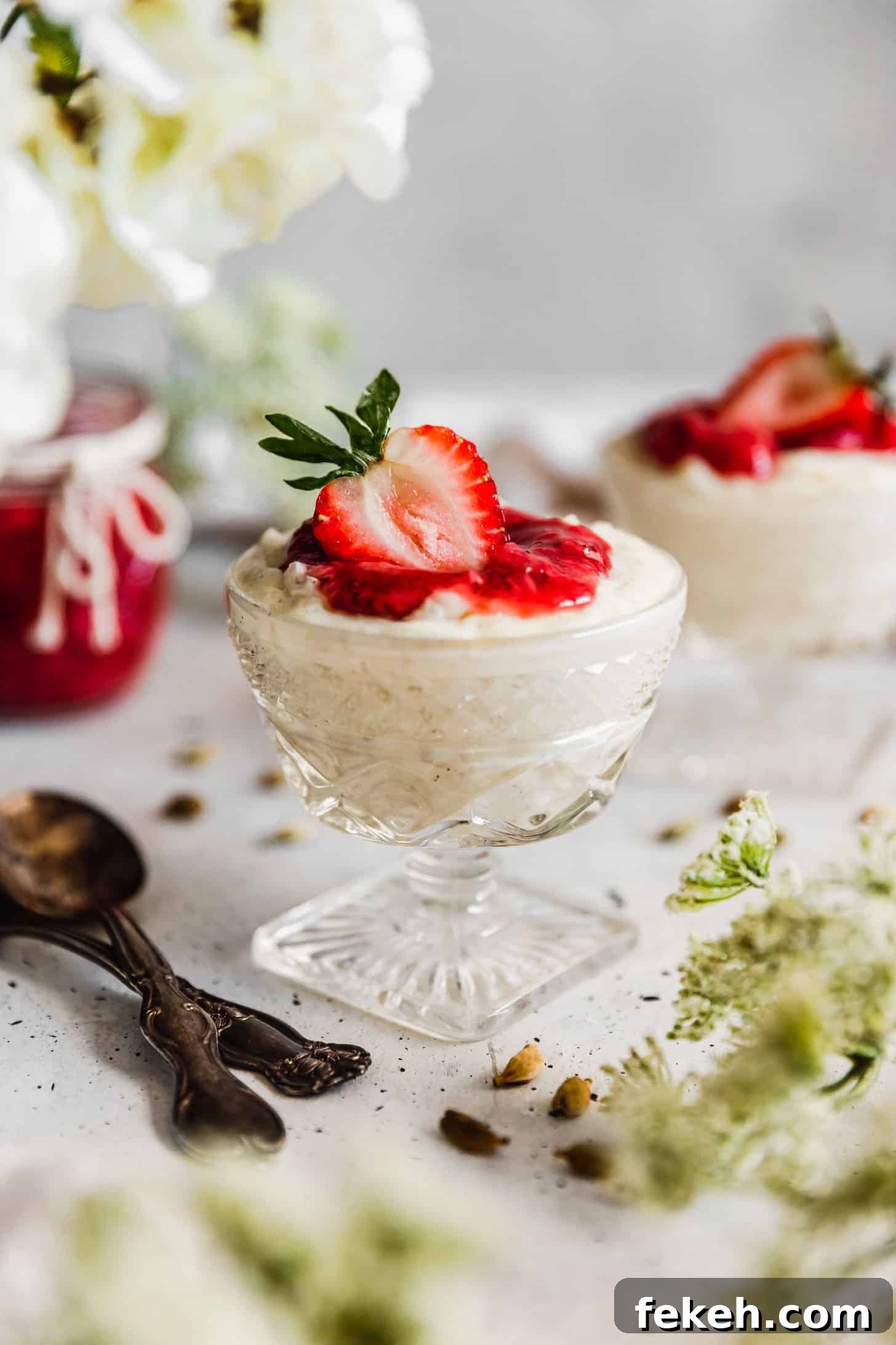 Two glass bowls of cardamom rice pudding with strawberry compote on a white counter next to a jar of jam, spoons, cardamom pods, and white blossoms.