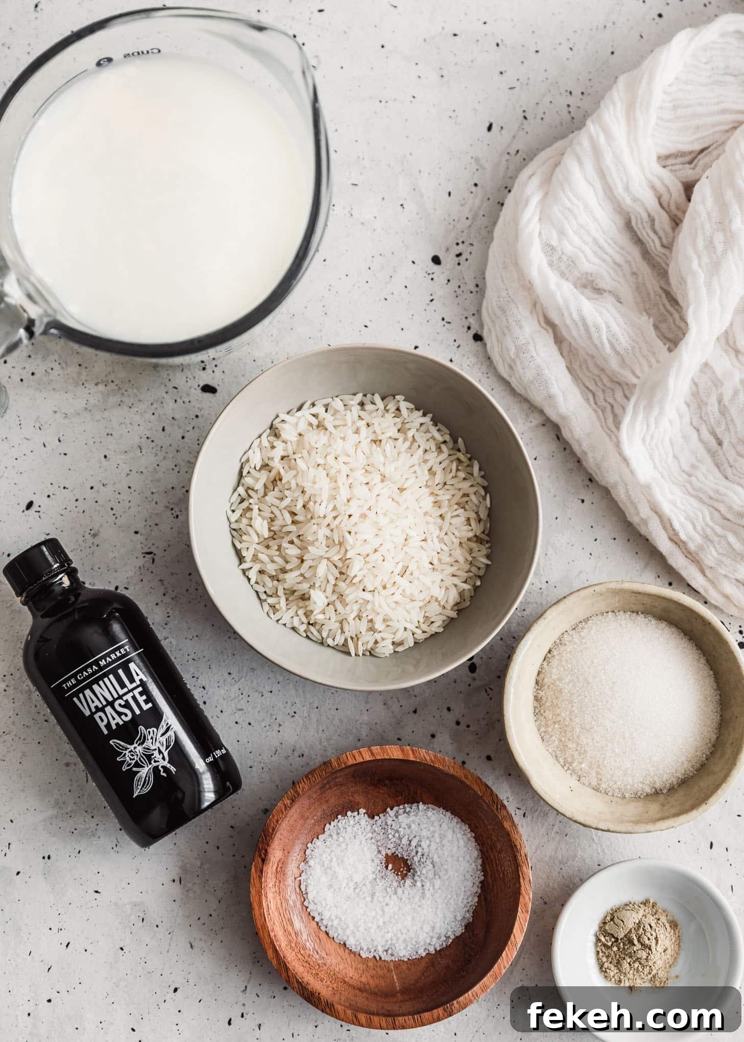 Wood and white bowls of rice, sugar, milk, cardamom, and salt on a white counter next to a bottle of vanilla bean paste.
