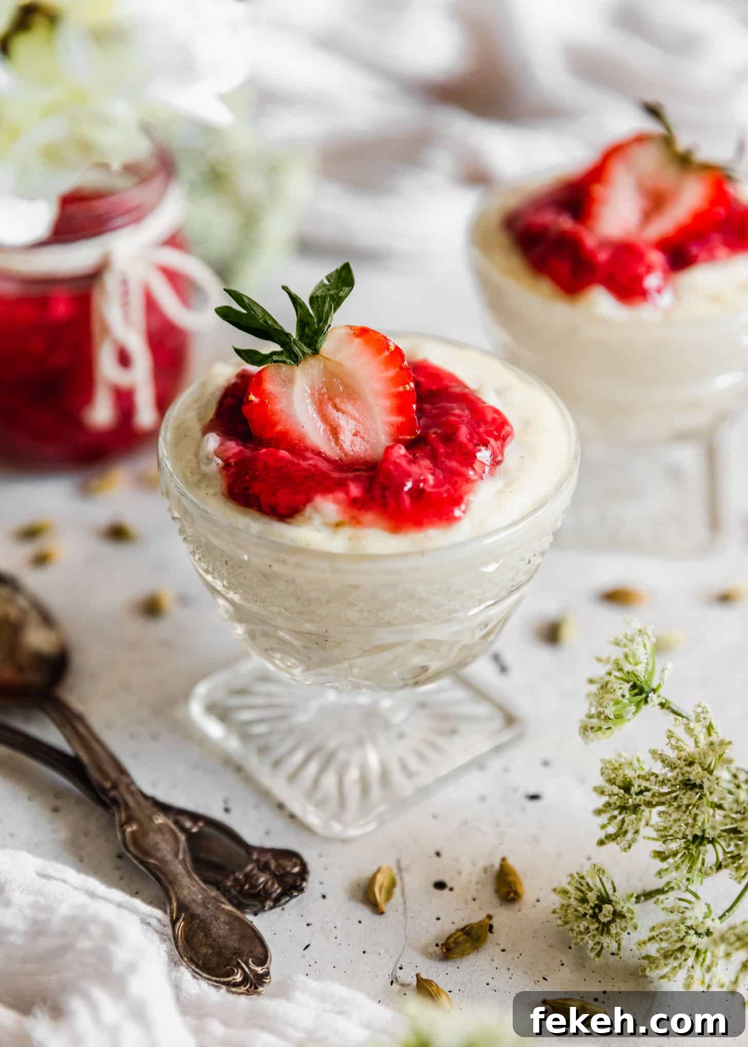 Two glass dishes of cardamom rice pudding with strawberries next to cardamom pods, a jar of jam, spoons, and white blossoms on a white counter.