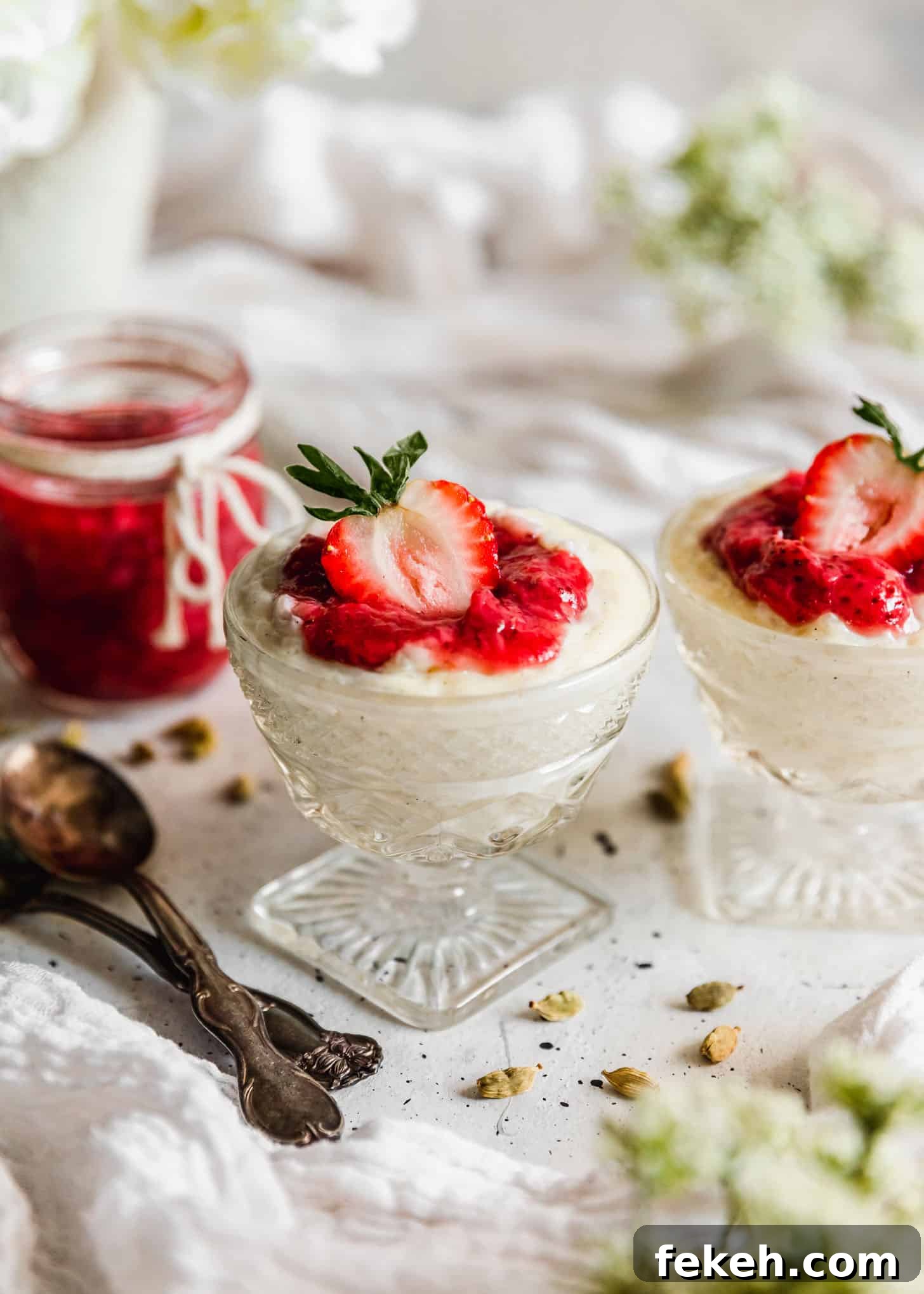 Two glass dishes of cardamom rice pudding with strawberries next to jam, spoons, cardamom pods, and white flowers on a white table.