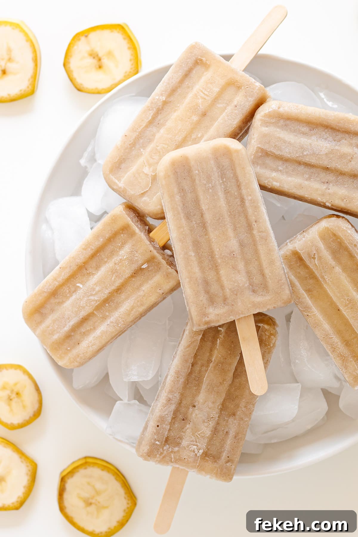 Homemade Banana Popsicles: Creamy No-Sugar Frozen Treats 2 Banana popsicles sitting on a round white platter of ice cubes with slices of banana beside it.