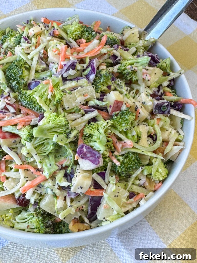Creamy Broccoli Slaw with Crunchy Apples and Almonds 3 Overhead view of a white bowl full of crunchy, creamy broccoli slaw with a silver serving spoon on a yellow napkin.