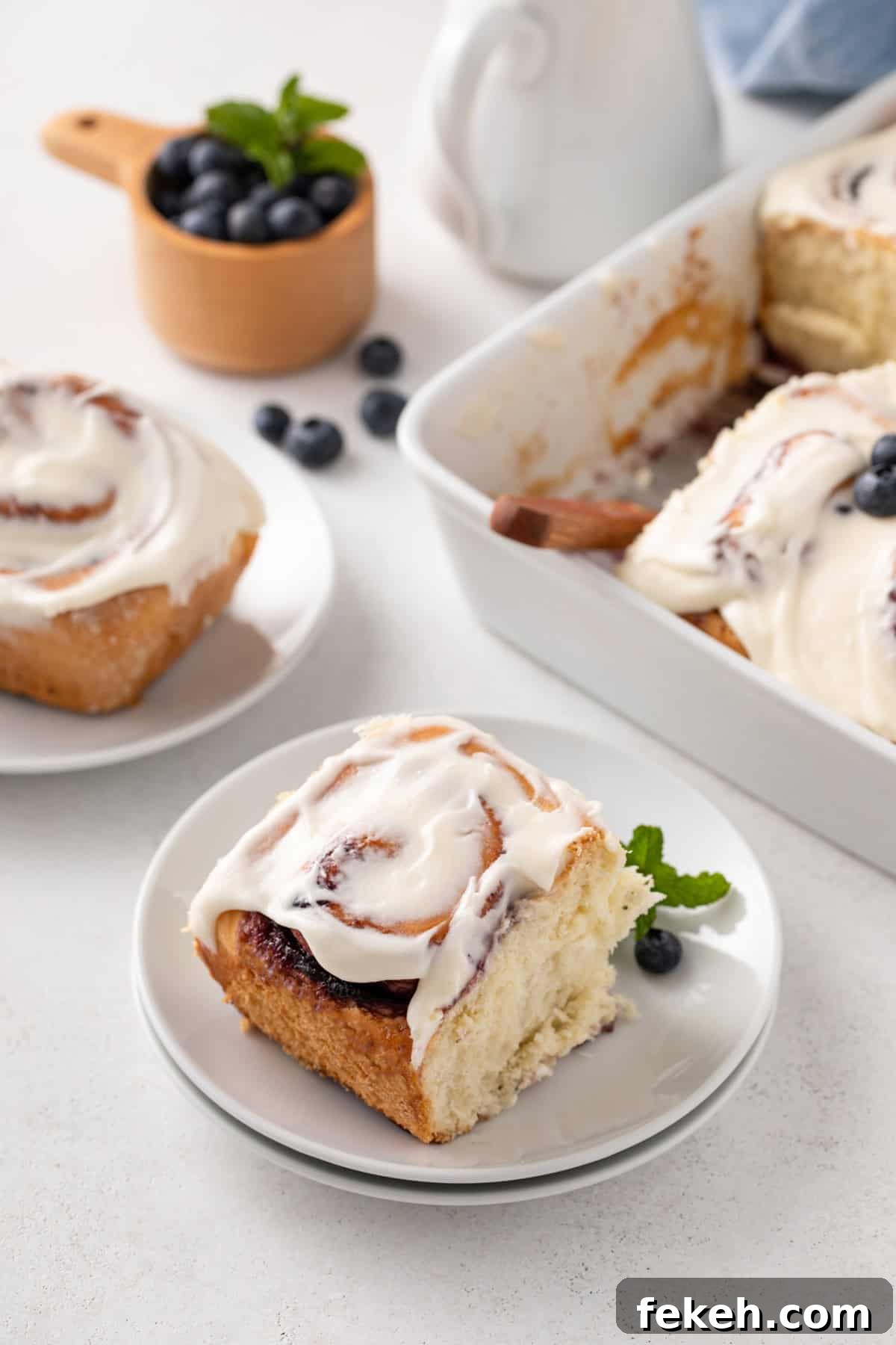 White plate holding a blueberry sweet roll, with a second plate and the pan of rolls in the background.