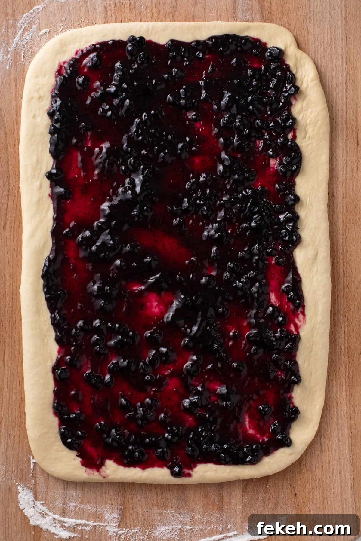 Sweet roll dough rolled into a rectangle on a wooden surface and spread with blueberry preserves.