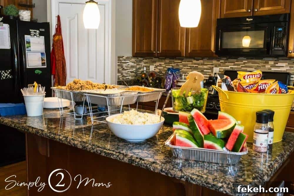 Kitchen island with trays of food, bucket of chip bags, and bowl of cut watermelon