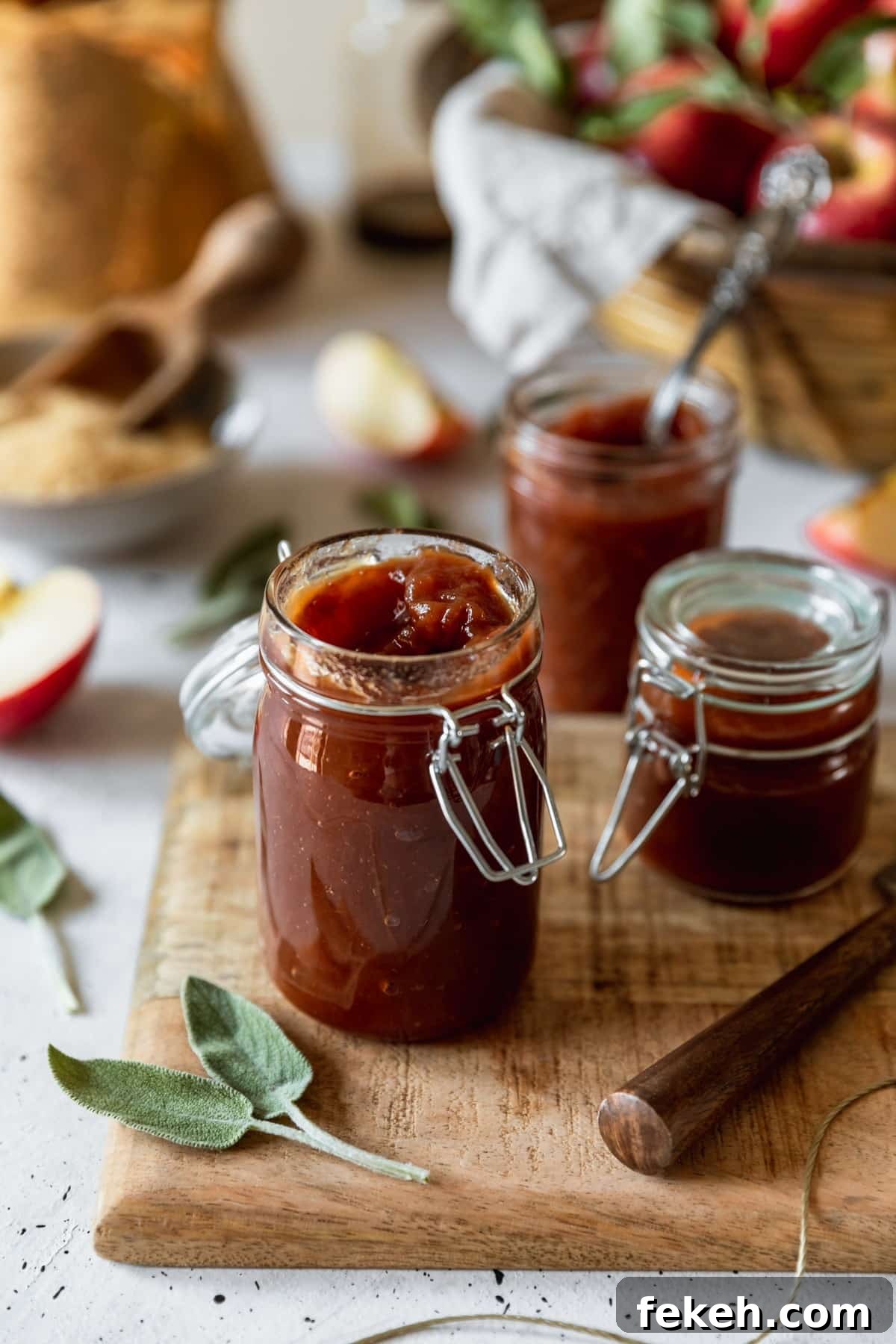 A closeup image of a jar of apple butter on a wood board placed on a white counter. In the background are two more jars of apple butter, a basket of apples, sliced apples, and sage leaves.