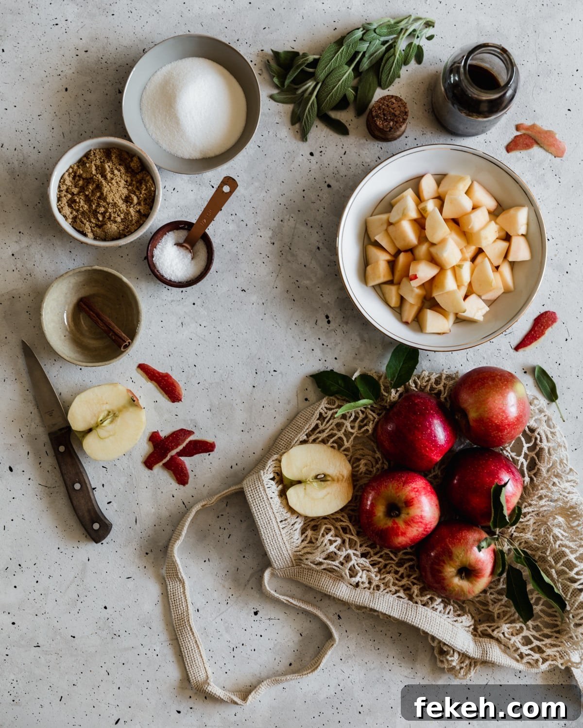 A mesh bag filled with apples on a grey counter next to a white bowl of diced apples, white and grey bowls with sugar, brown sugar, and cinnamon, a bunch of sage, and a bottle of vanilla extract.