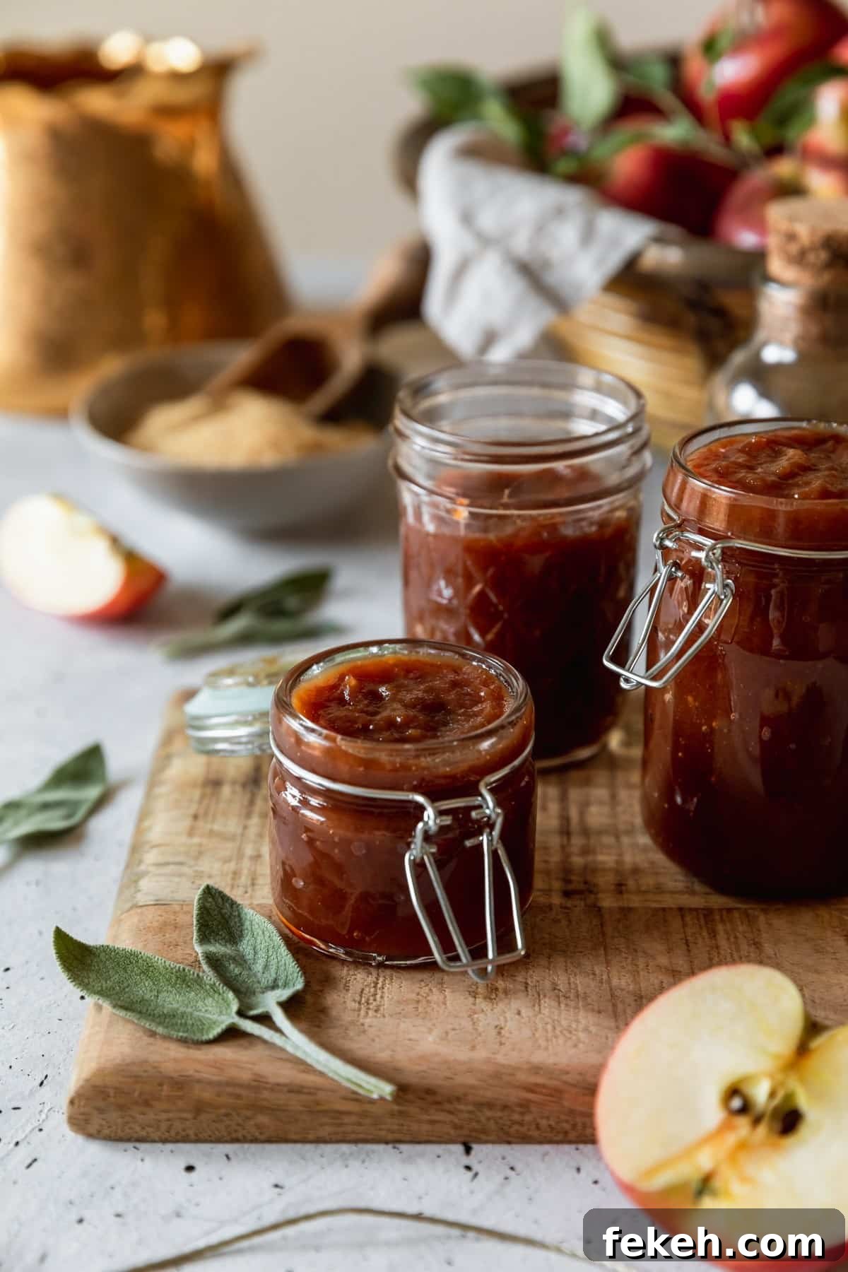 A closeup side image of a small jar of apple butter with two larger jars on the background on a wood board with sage leaves. The board is on a white speckled table with a gold teapot, basket of apples, and grey bowl of sugar in the background.