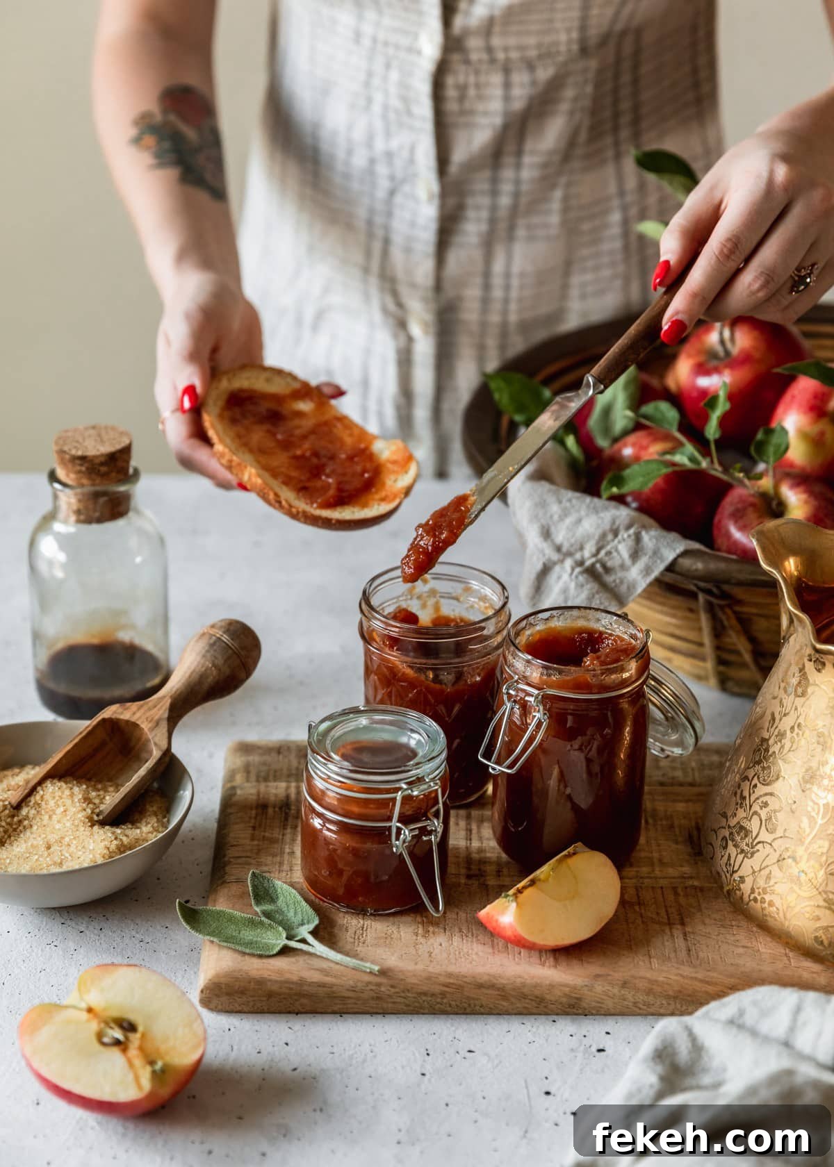 A side image of three jars of apple butter on a wood board next to a gold teapot. In the background is a basket of apples and a woman wearing a white and grey dress spreading apple butter on a slice of toast.