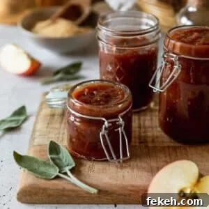 A side image of a small jar of apple butter on a wood board with two more jars in the background. the board is on a white counter with sliced apples, sage leaves, and a bowl of sugar in the background.