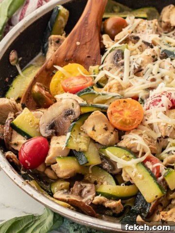 Cooked chicken, zucchini, tomatoes, and mushrooms in metal pan with wooden spoon.