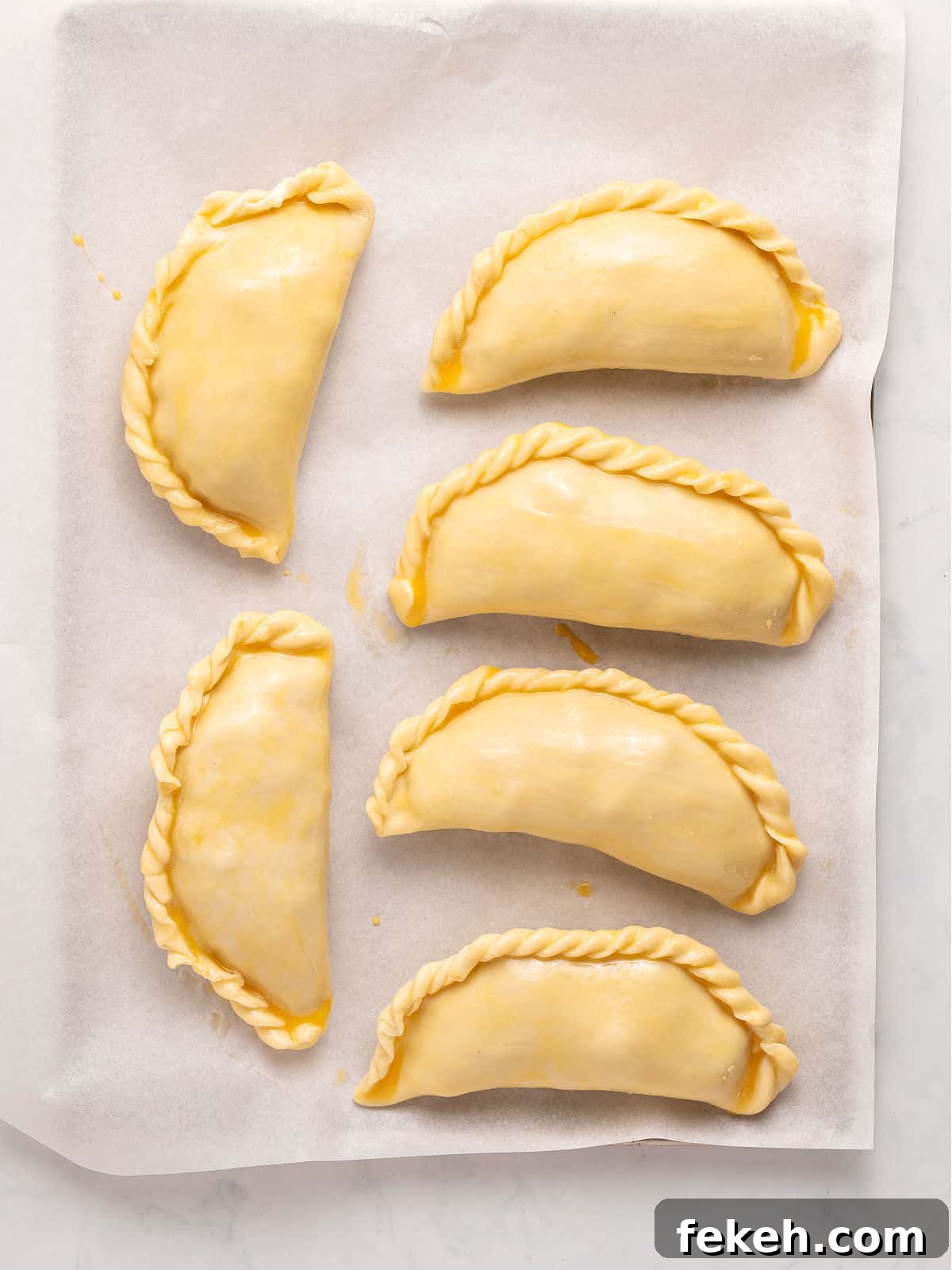 Knife-Cut Beef Empanadas: Crispy Handheld Meat Pastries 9 Six empanadas with egg wash before baking on white parchment paper.