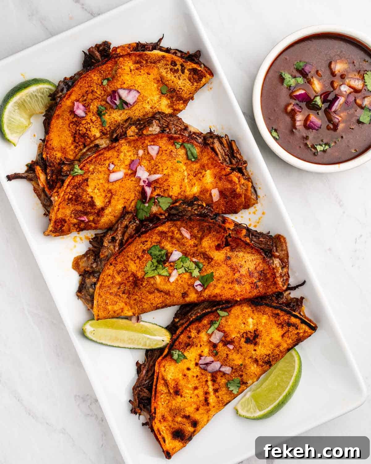 Authentic Quesabirria Tacos: How to Make Birria Tacos at Home 3 Four birria tacos on a plate with lime wedges and consommé dipping sauce.