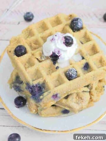 Three blueberry protein waffles stacked on a white plate topped with whip cream, blueberries and icing sugar.