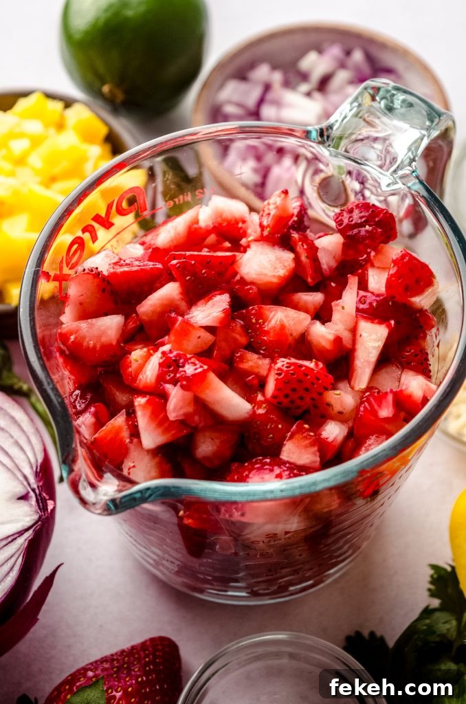 Fresh Strawberry Salsa Recipe: Zesty Fruit Salsa for Chips & Tacos 4 A measuring cup of chopped strawberries surrounded by other salsa ingredients like a red onion, yellow pepper, cilantro, and a lime.