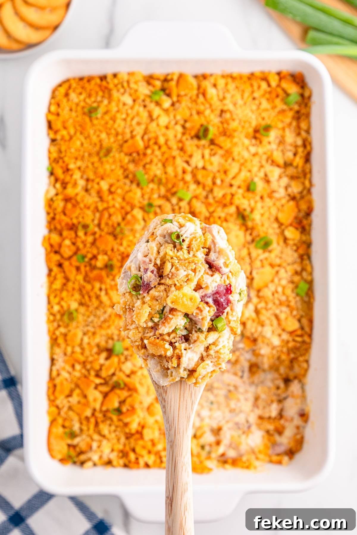 A wooden spoon holds a creamy casserole with a crunchy topping above a casserole dish.