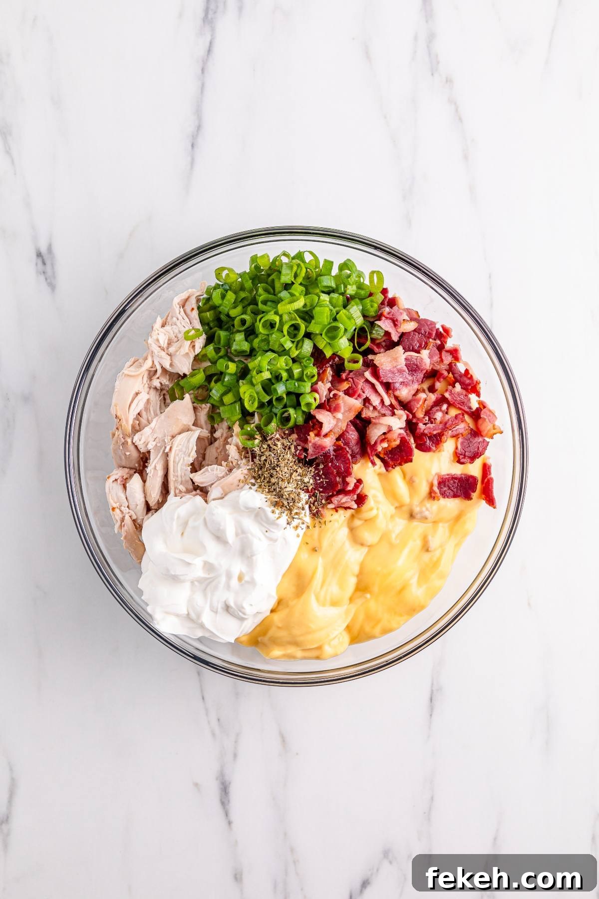 A bowl with shredded chicken, chopped green onions, bacon, sour cream, soup, and seasonings on a marble surface.