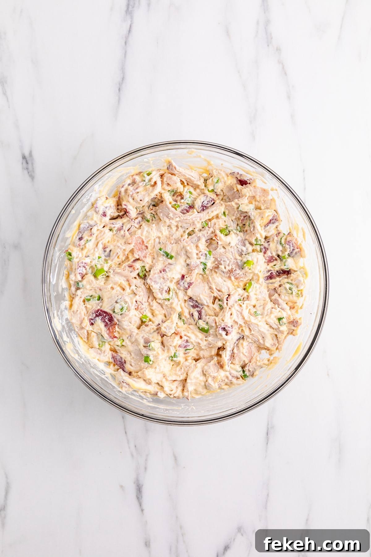 A glass bowl of creamy dip with chopped green onions and pieces of bacon on a marble surface.