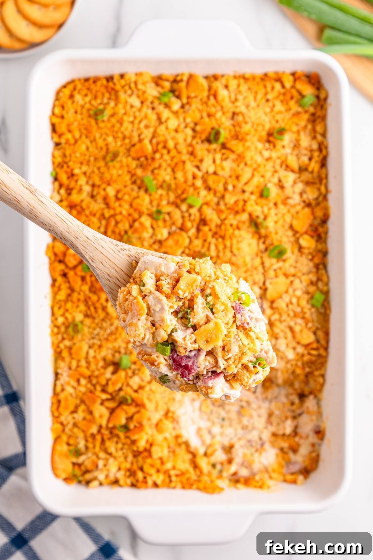 A wooden spoon holds cheesy casserole with a crunchy topping over a white baking dish of the same casserole.