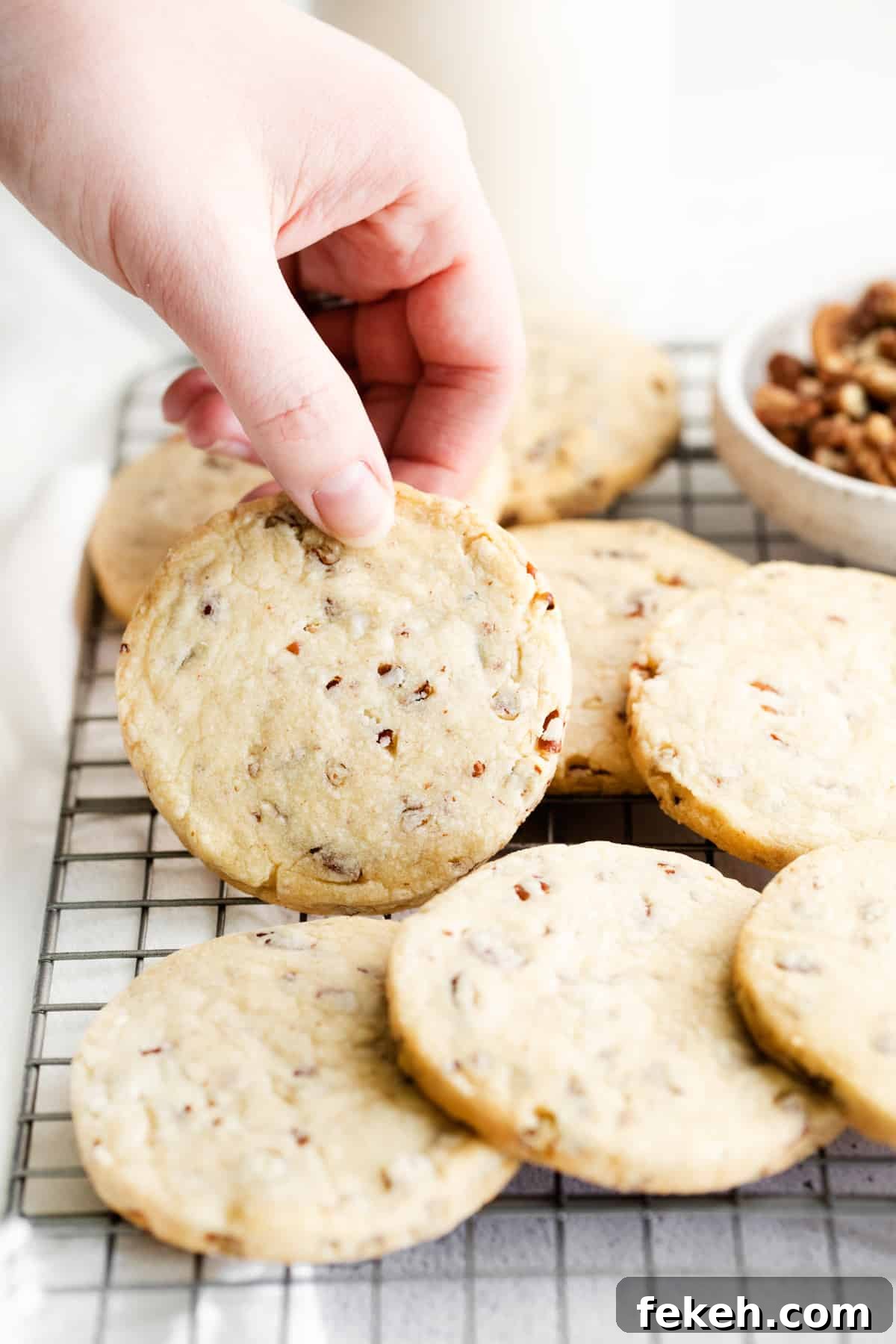 a hand holding a pecan shortbread cookie upright to show the top, next to a pile of cookies on a wire rack.