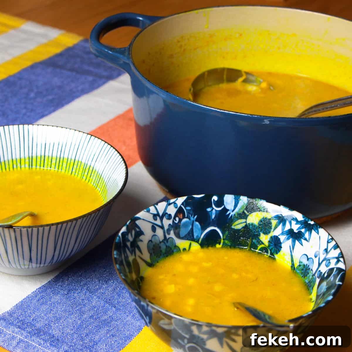 a big navy le creuset pot full of spiced pumpkin and sweetcorn soup with two bowls of soup in the foreground. All on a stripy blue, yellow, orange and white tablecloth on a wooden table.