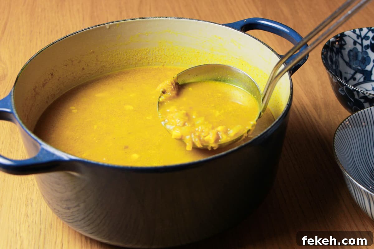 Navy casserole pan full of spiced pumpkin and sweetcorn soup and a ladle taking a scoop out of the pan to go in the two blue and white decorated bowls next to it.  All on a wooden table.