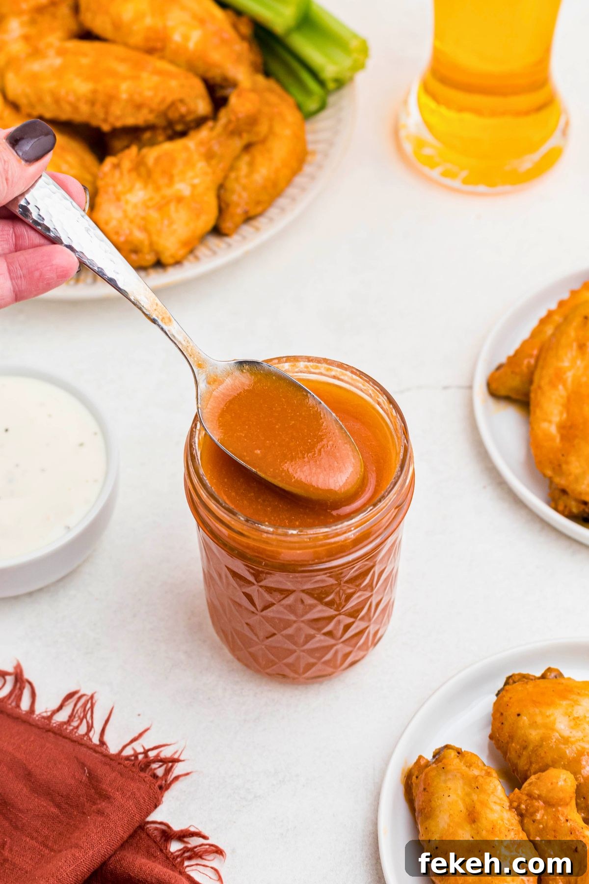 Orange sauce in a small jar, being spooned out, in front of plates of chicken wings. 