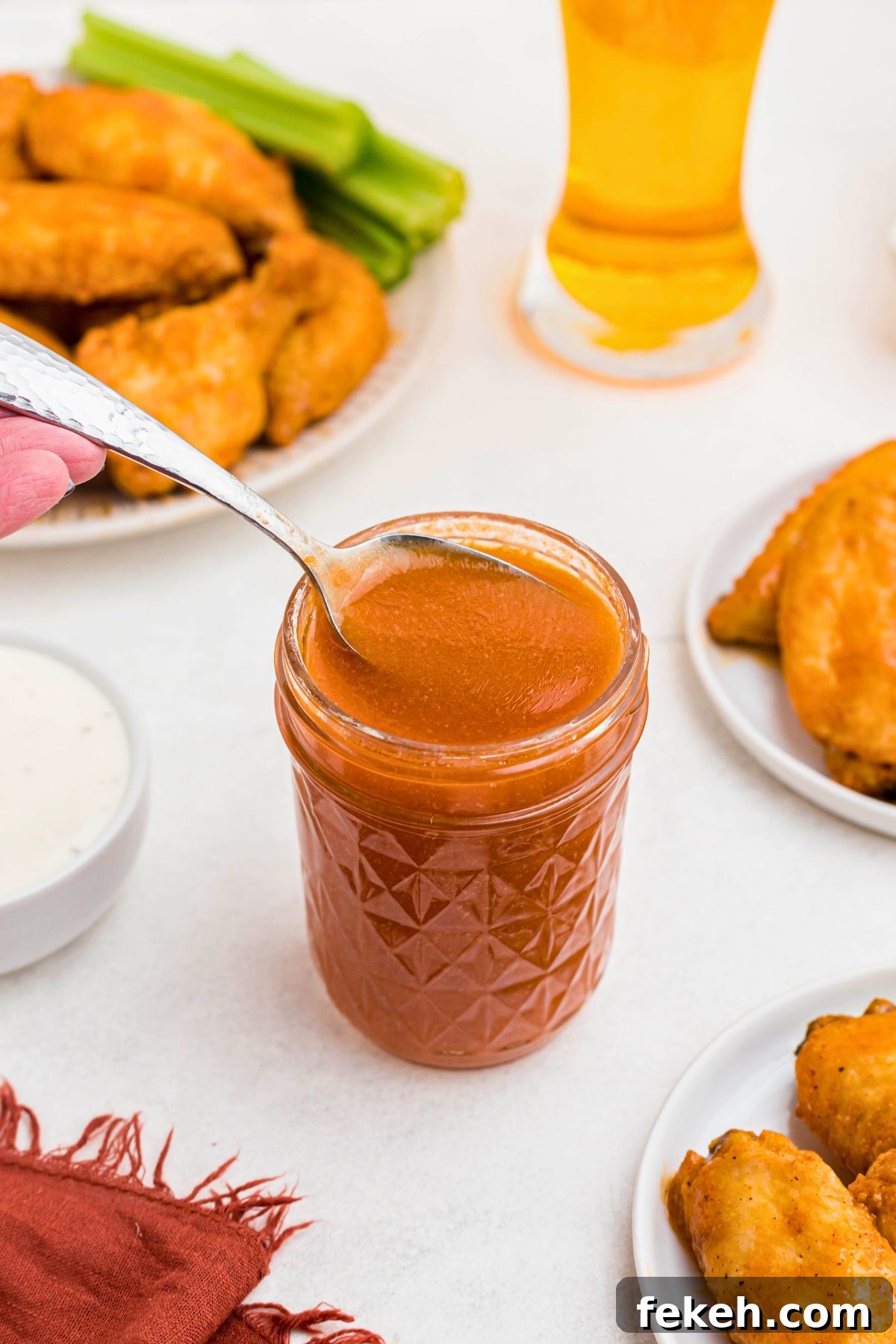 Orange sauce in a small jar, being spooned out, in front of plates of chicken wings.